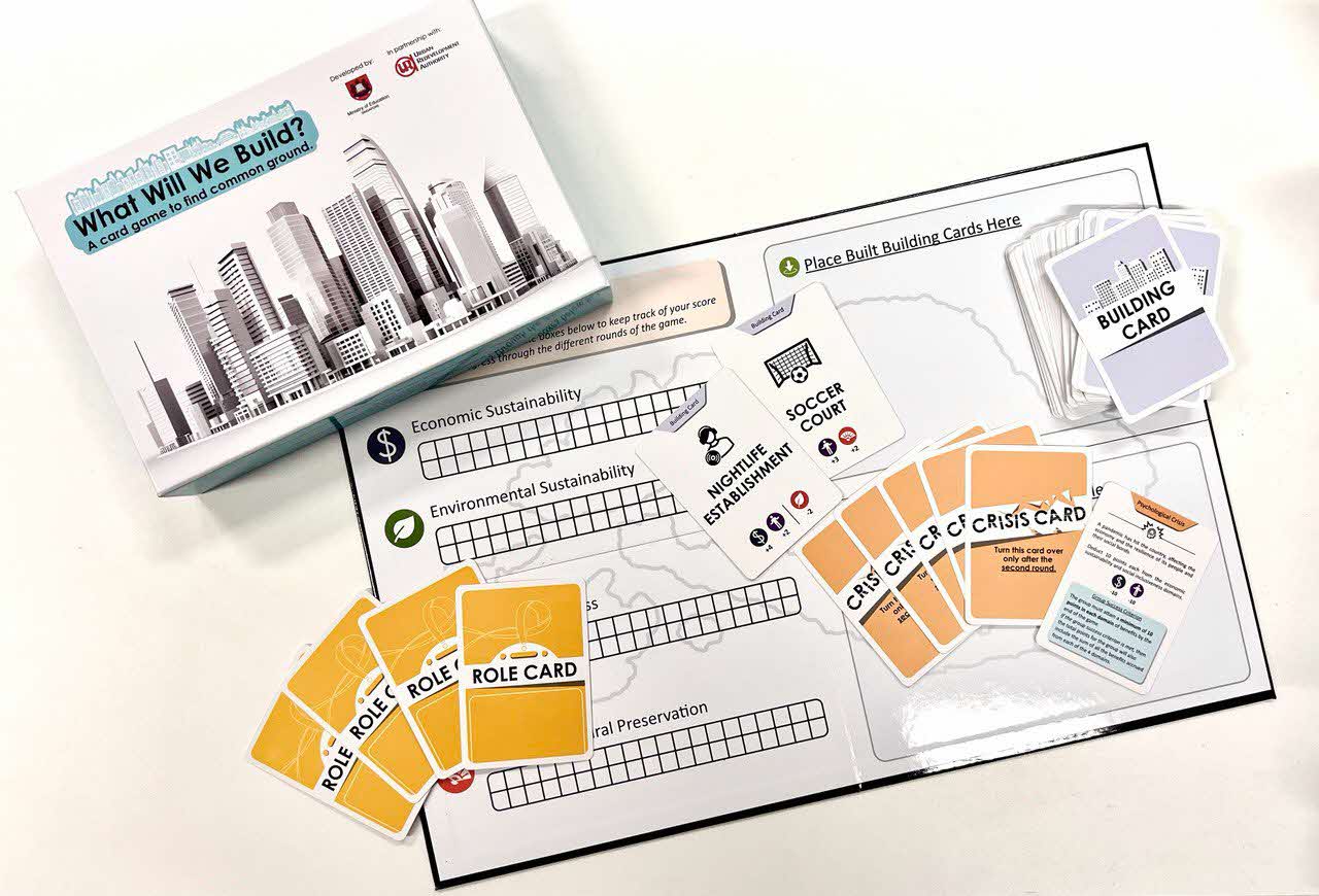 Board game set with “What Will We Build?” box, featuring building, role, and crisis cards arranged on a game board with sustainability score tracks.